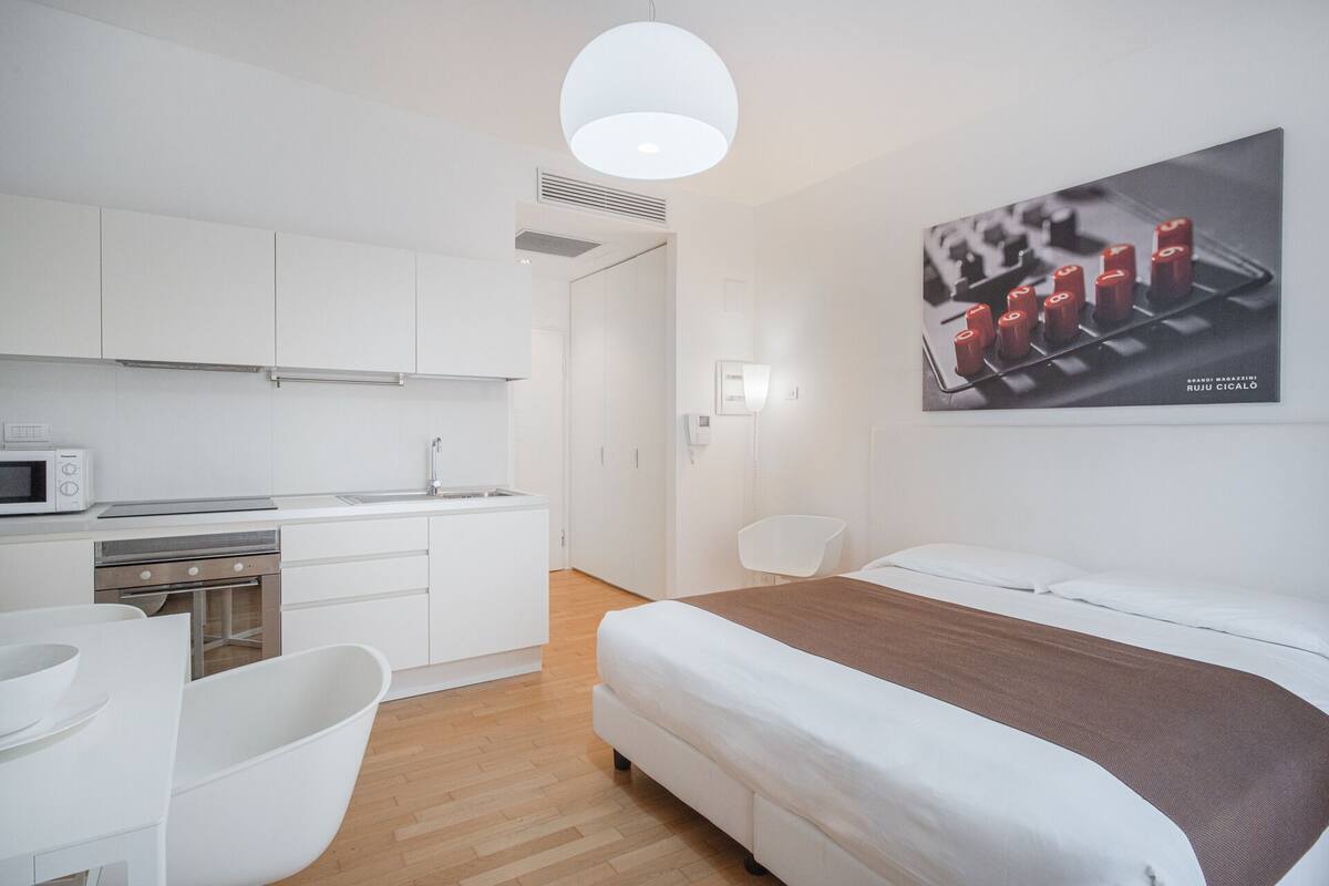 A modern hotel room with a kitchenette, a bed, a white chair, and a wall-mounted artwork.
