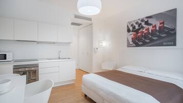 A modern hotel room with a kitchenette, a bed, a white chair, and a wall-mounted artwork.