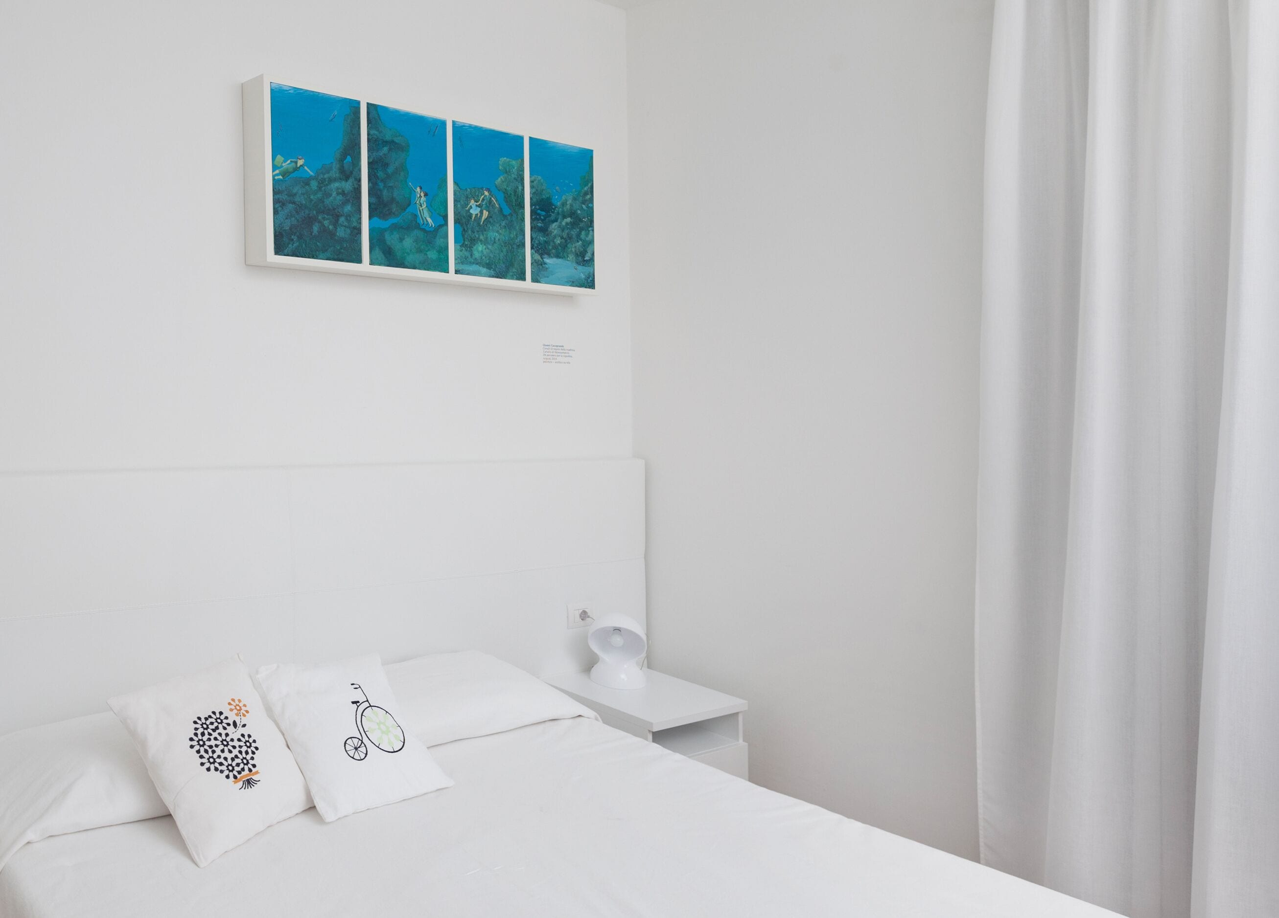 Habitación de hotel minimalista con una cama grande, ropa de cama blanca, una mesita de noche con lámpara y un cuadro en la pared que representa una escena submarina.