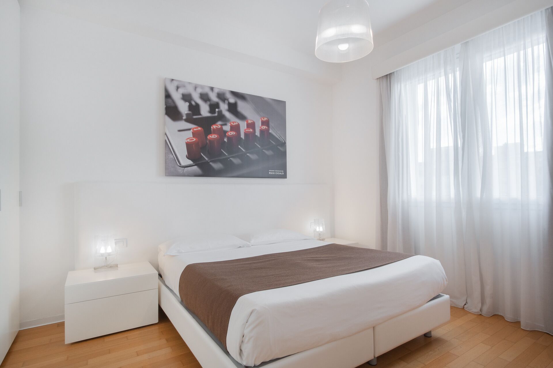 A modern bedroom with a bed, white nightstands, a wall-mounted artwork, and sheer curtains.