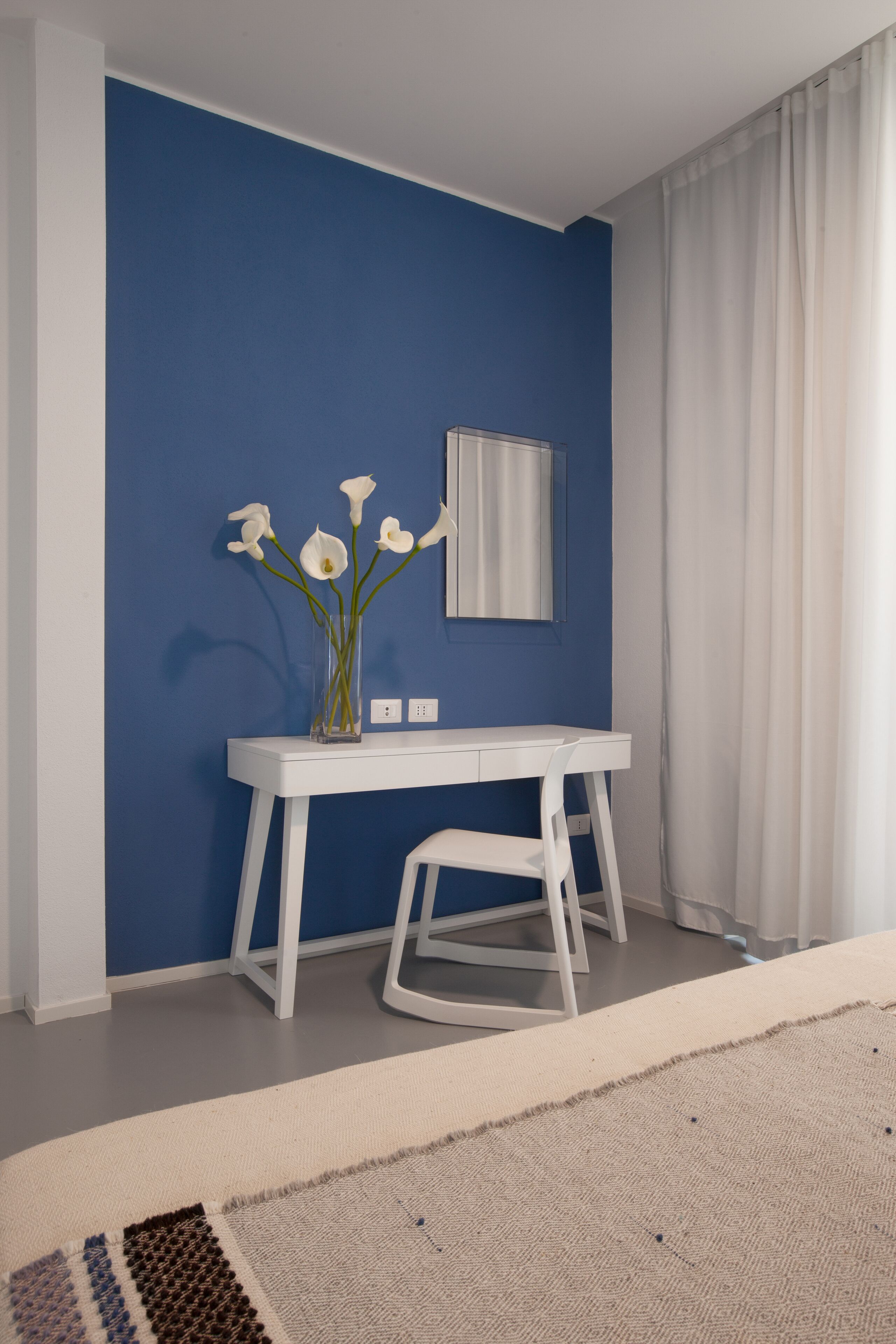 A room with a blue wall, a white desk with a vase of flowers, a chair, and a bed with a beige rug.
