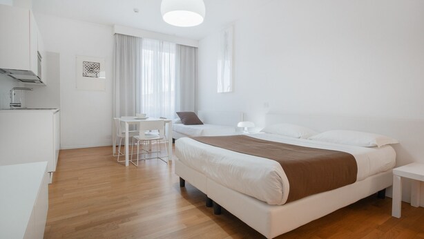 A modern, minimalist bedroom with a large bed, bedside tables, a small desk, and a dining area. - Felix Hotels - Residence Grandi Magazzini (Nuoro)