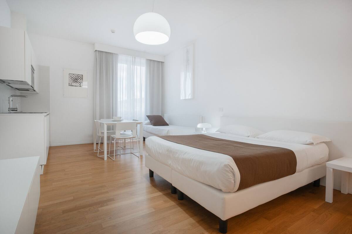 A modern, minimalist bedroom with a large bed, bedside tables, a small desk, and a dining area.