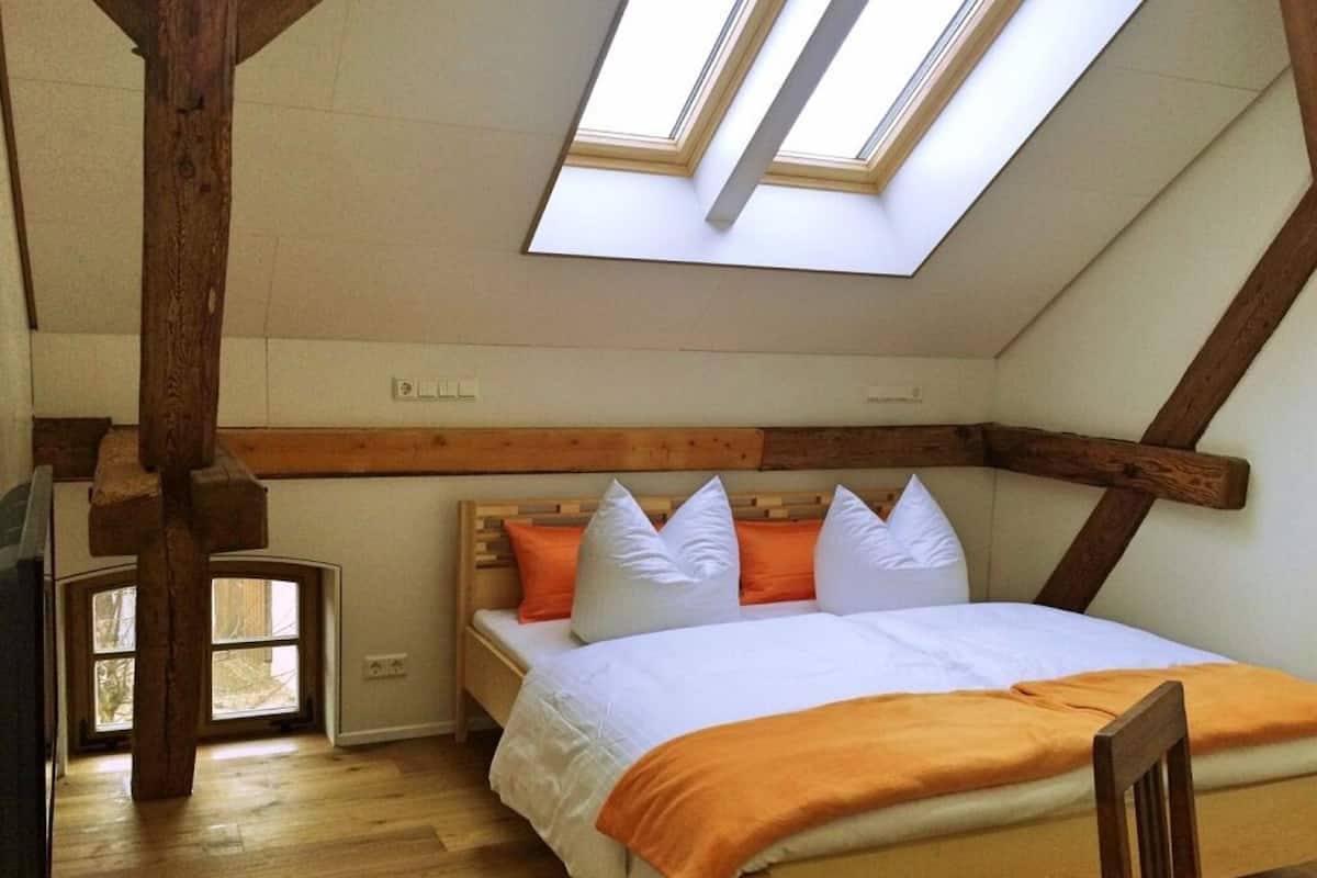 A bedroom with a sloped ceiling, wooden beams, a bed with white and orange bedding, and skylights.