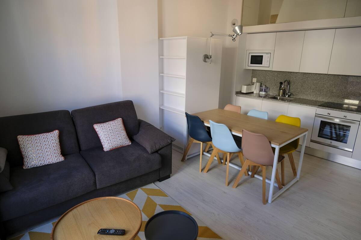 Una sala de estar moderna con un sofá gris oscuro, una mesa de centro de madera y una zona de comedor con una mesa de madera y sillas coloridas.