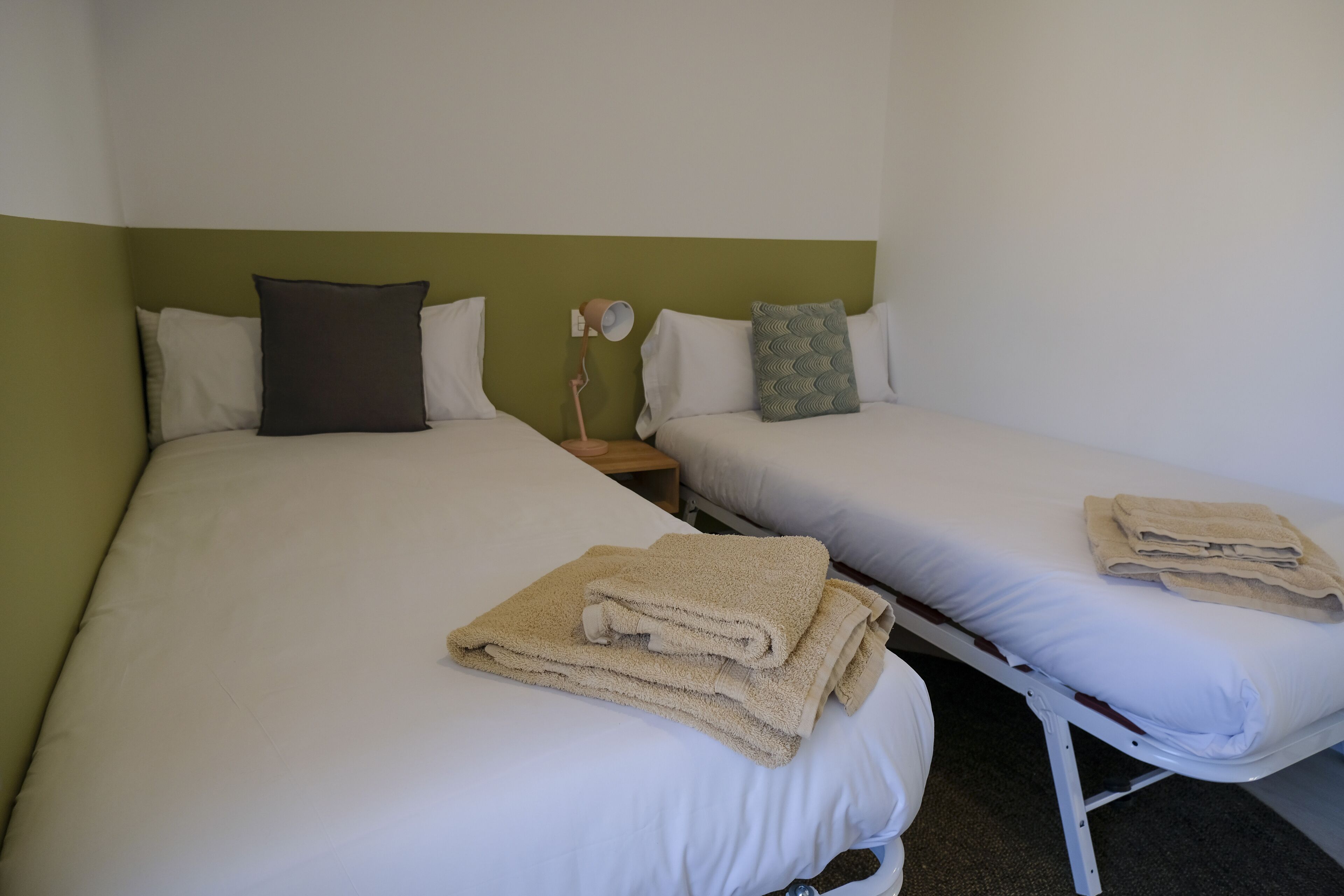 Una habitación con dos camas, cada una con ropa blanca y una almohada. Hay una mesita de noche con una lámpara entre las camas.