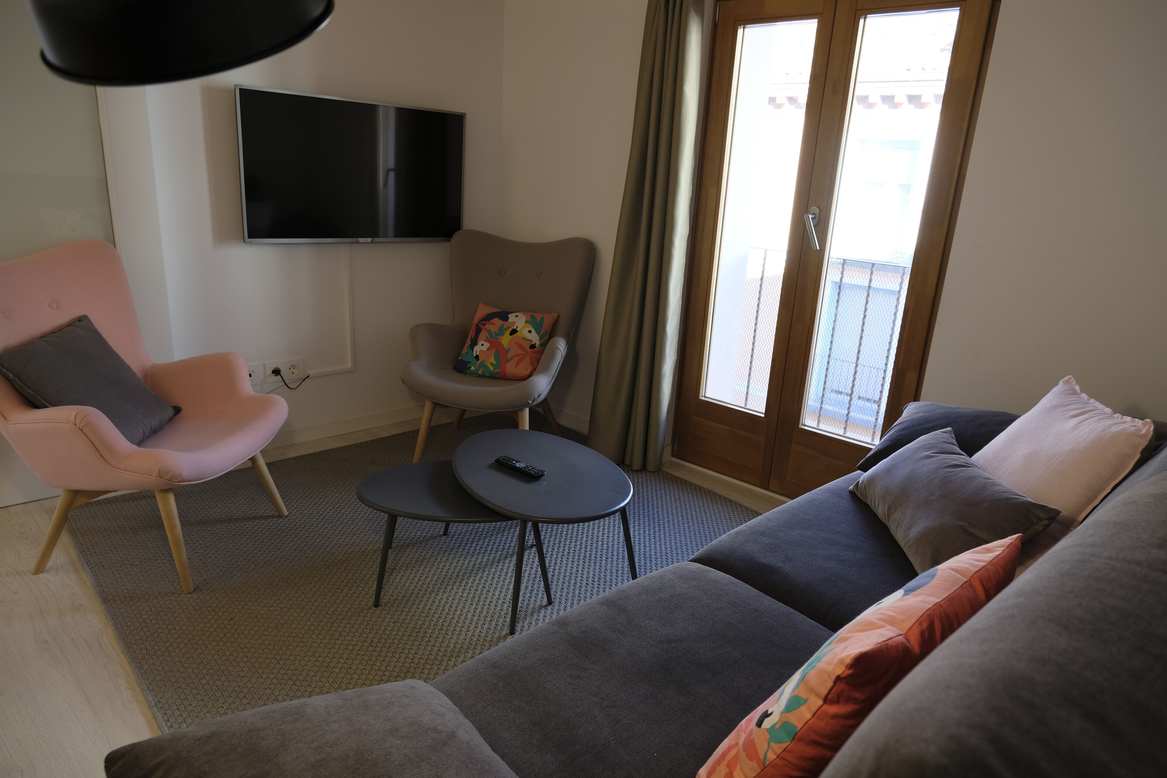 A living room with a grey sofa, a pink armchair, a small round table with a remote control, and a TV mounted on the wall.