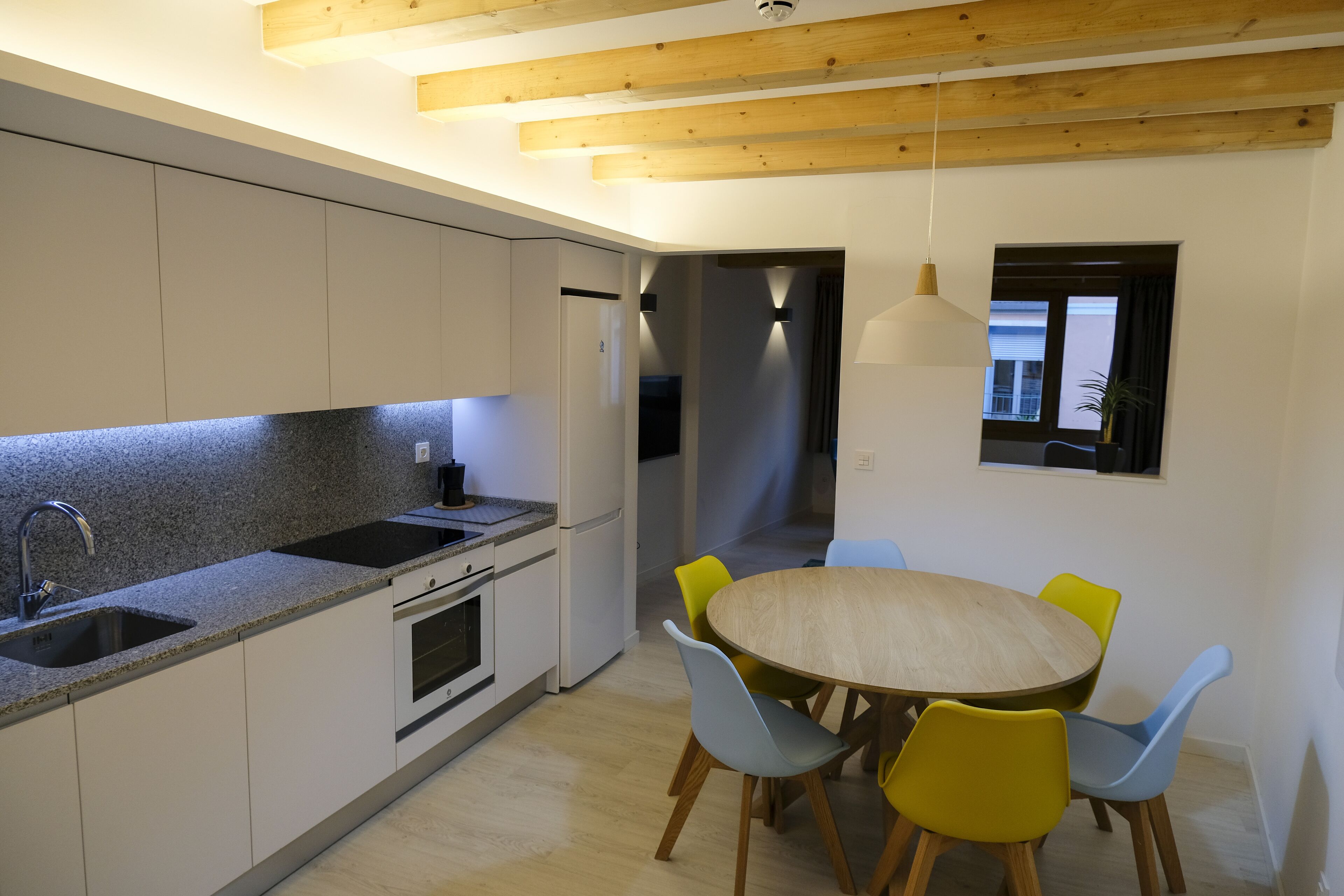 Une cuisine moderne avec des armoires blanches, un comptoir en granit et un four encastré. On y trouve un coin repas avec une table ronde en bois et quatre chaises, dont deux jaunes et deux bleues.