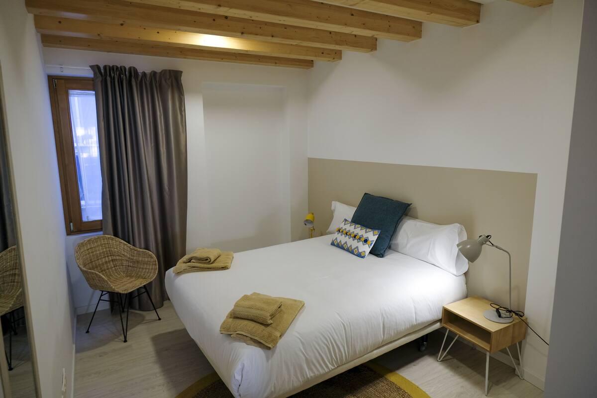 Un dormitorio con cama, una mesita de noche con lámpara, una silla y una ventana con cortinas.
