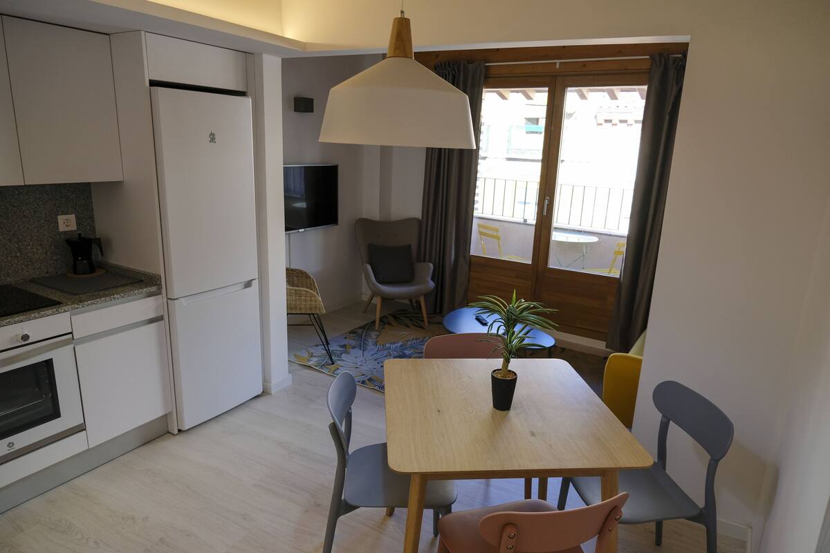 Cocina compacta con refrigerador, horno y cafetera. Hay un comedor con mesa, sillas y una planta en maceta. Un televisor está montado en la pared.