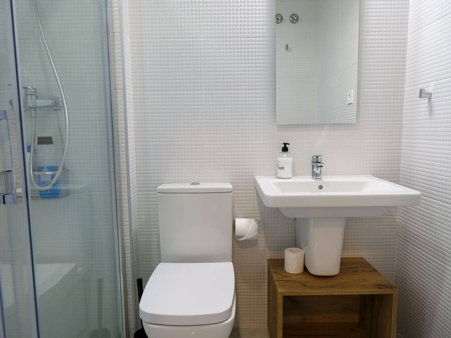 A modern bathroom with a glass shower enclosure, white tiled walls, a white toilet, a sink with a mirror above it, and a wooden shelf.