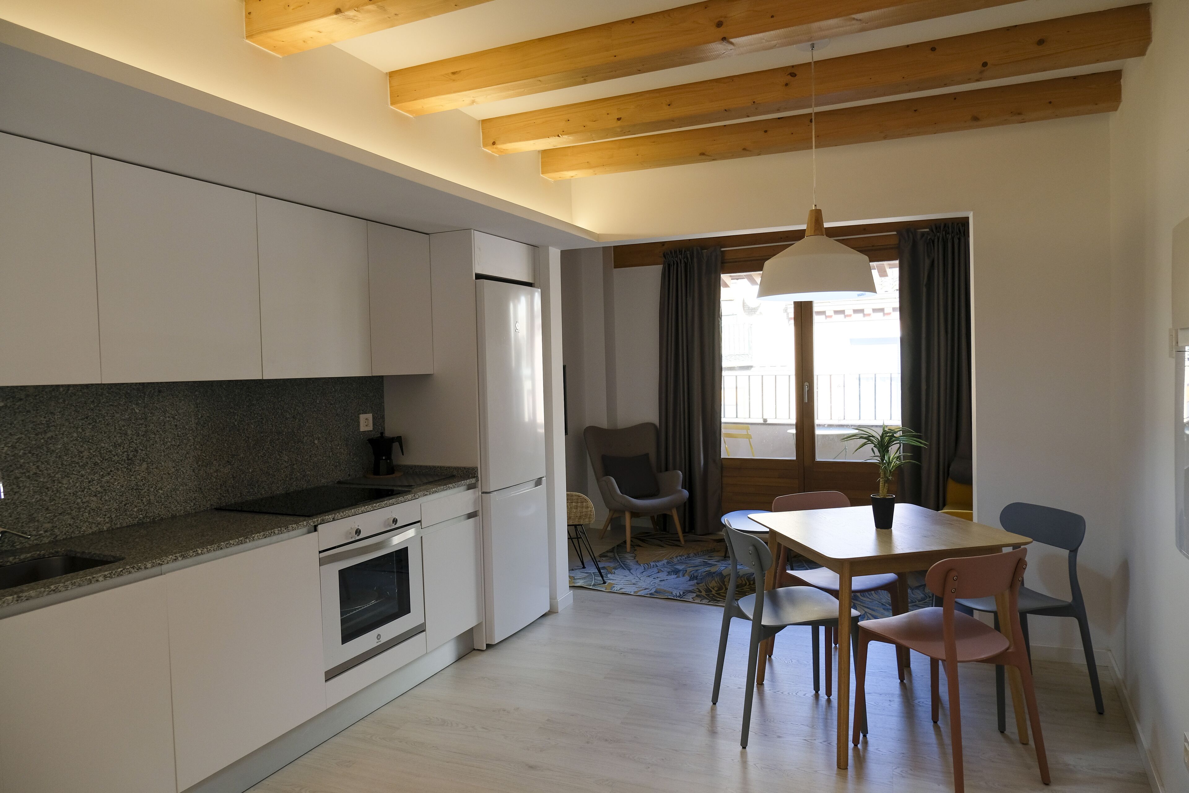 Una cocina moderna con armarios blancos, horno empotrado y encimera de granito. Hay una zona de comedor con mesa y sillas de madera, y una planta en maceta sobre la mesa.