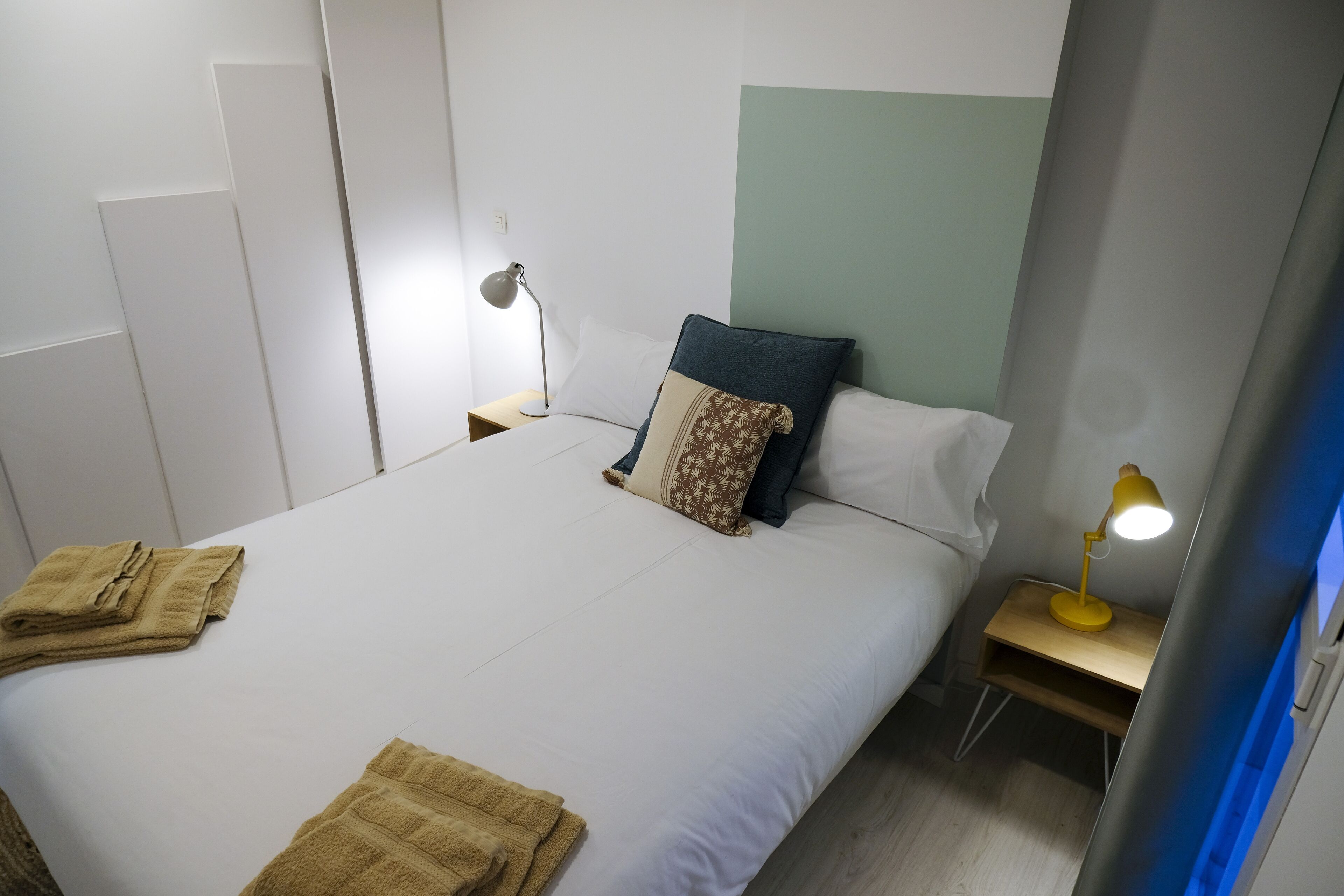 A neatly made bed with white linens, two pillows, a bedside lamp, and folded towels on the side.