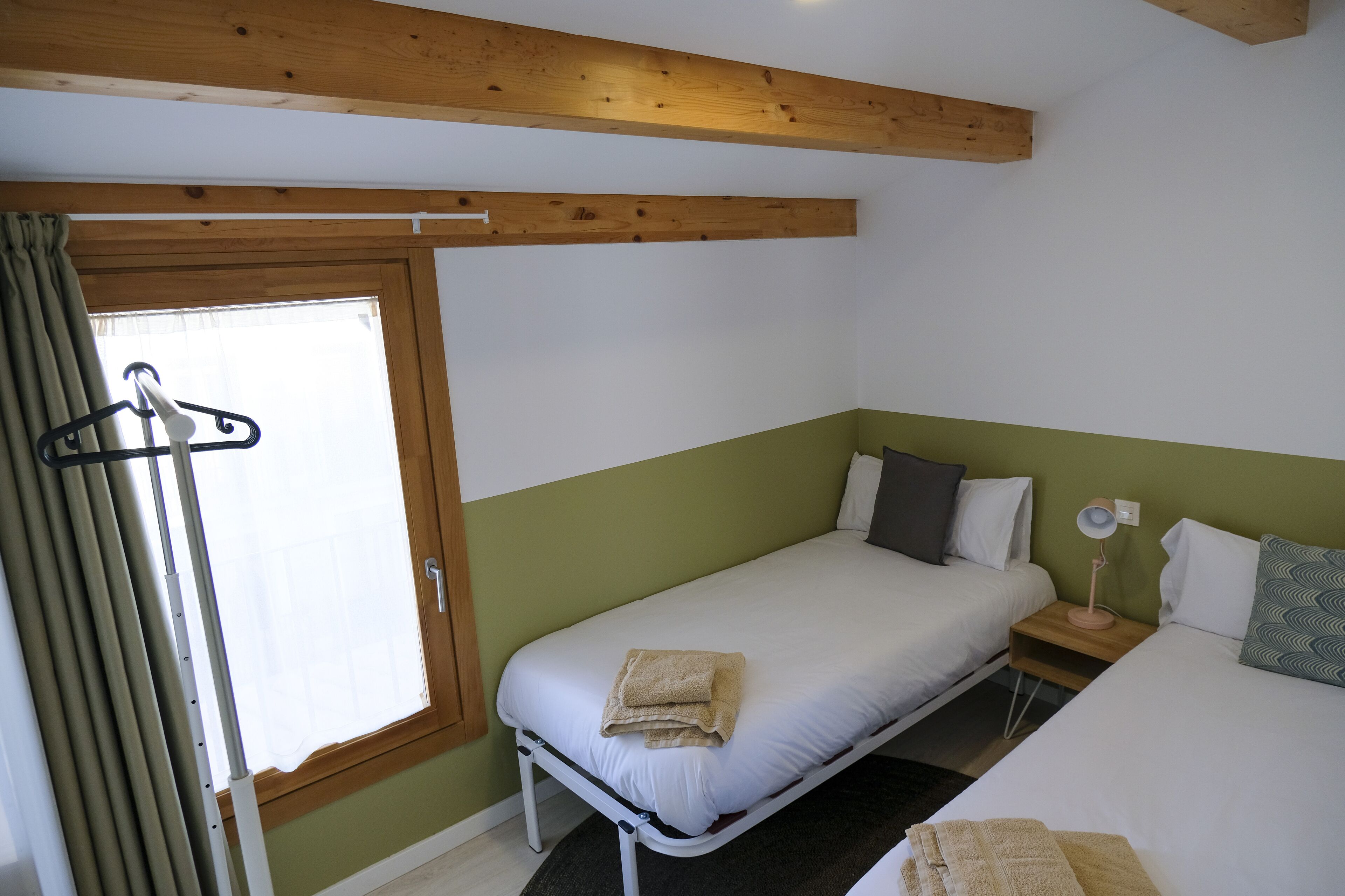Una cama individual con colchón blanco, cabecera de madera, mesita de noche con lámpara y ventana con cortinas.
