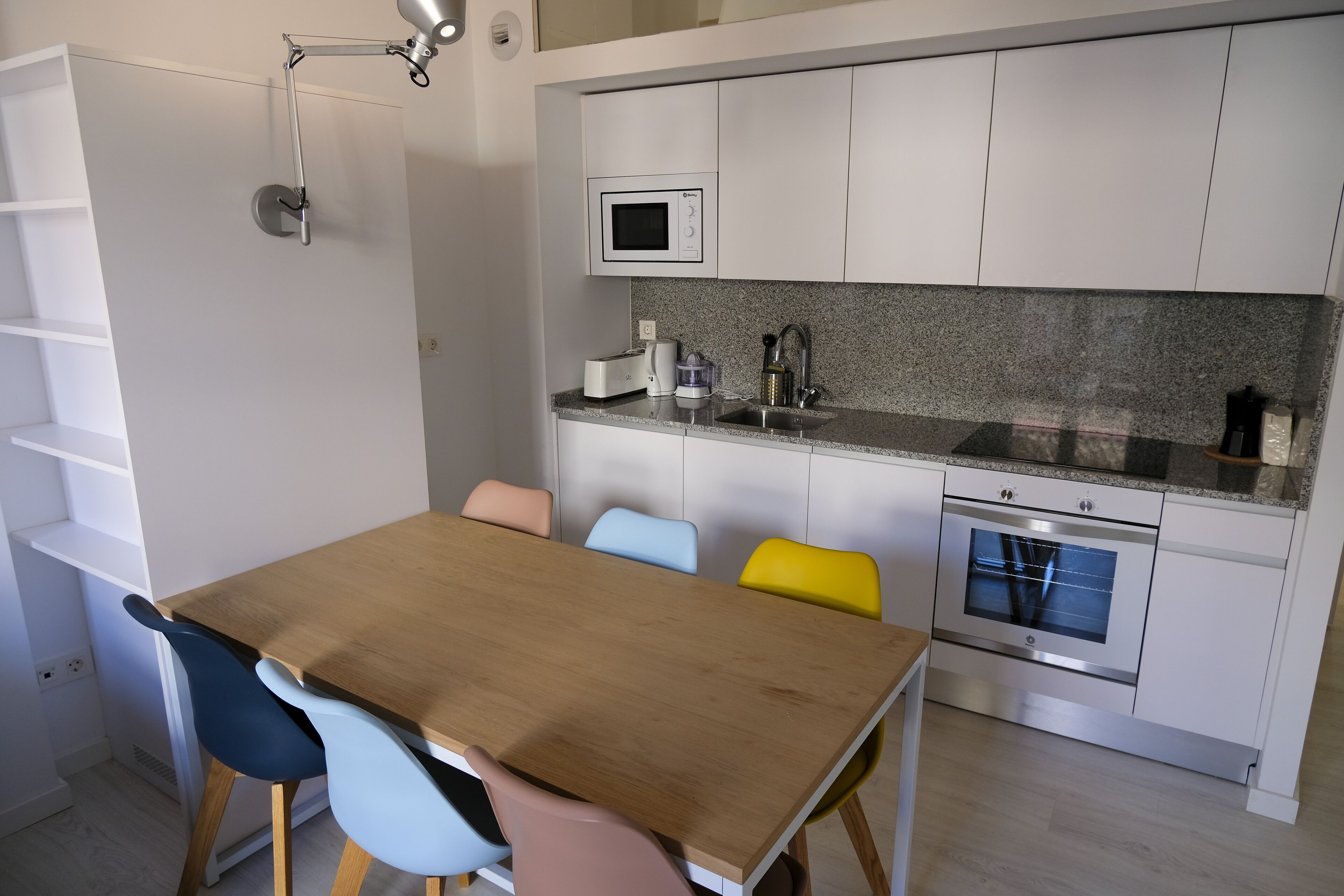 Une cuisine moderne avec des meubles blancs, un four à micro-ondes, un évier et une table à manger en bois, assortie de chaises colorées.