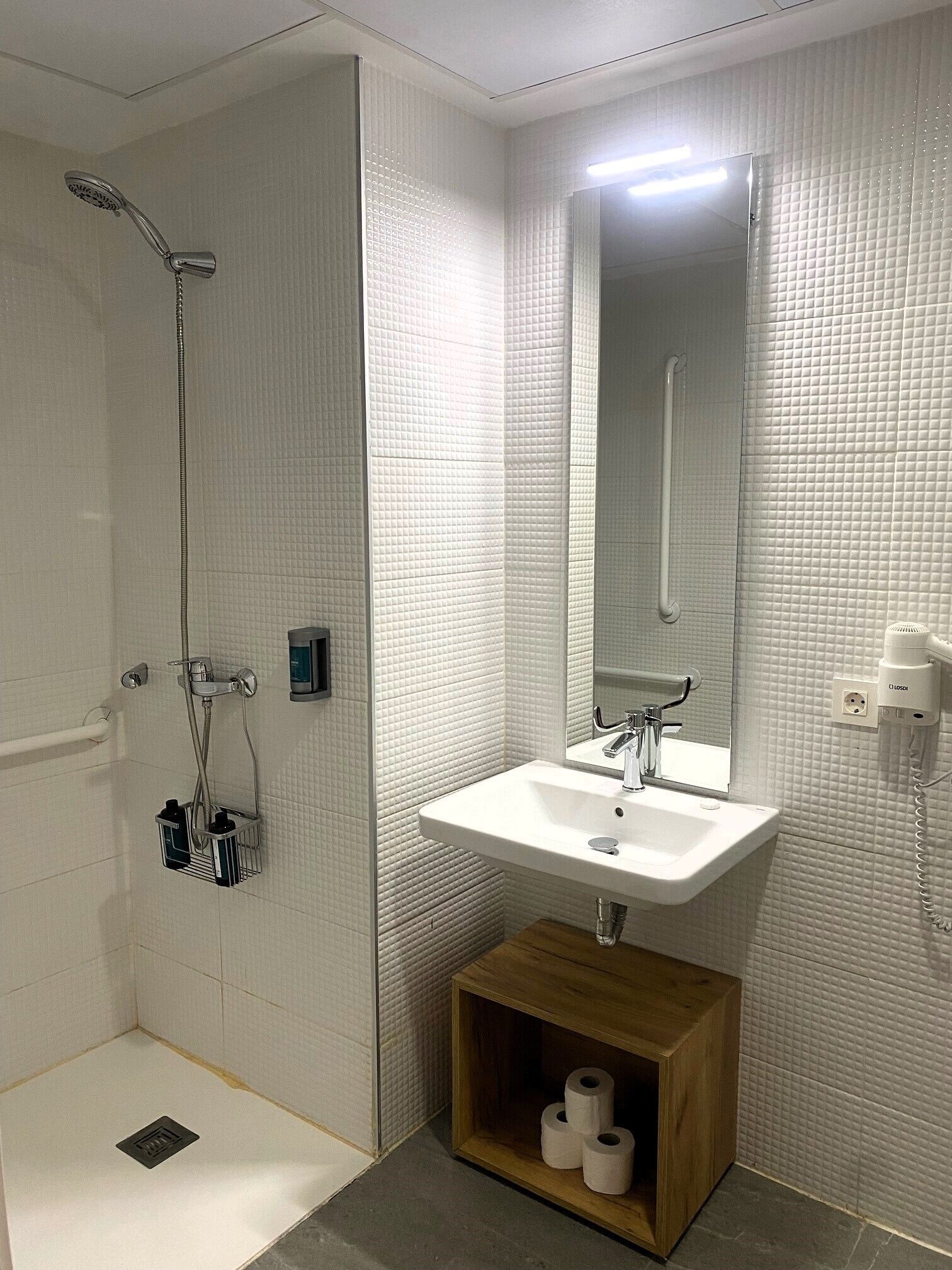 Une salle de bain équipée d’une douche, d’un lavabo, d’un miroir et d’un espace de rangement pour le papier toilette.
