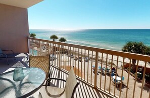 A balcony with a glass table and chairs, overlooking a beach with a clear view of the ocean.
