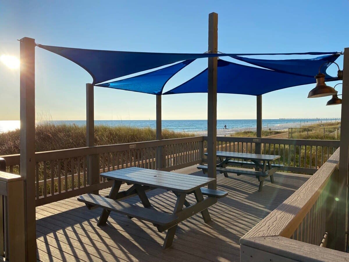 Terraza de madera con mesas de picnic y un toldo azul con vistas a la playa.
