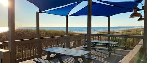 Terraza de madera con mesas de picnic y un toldo azul con vistas a la playa.