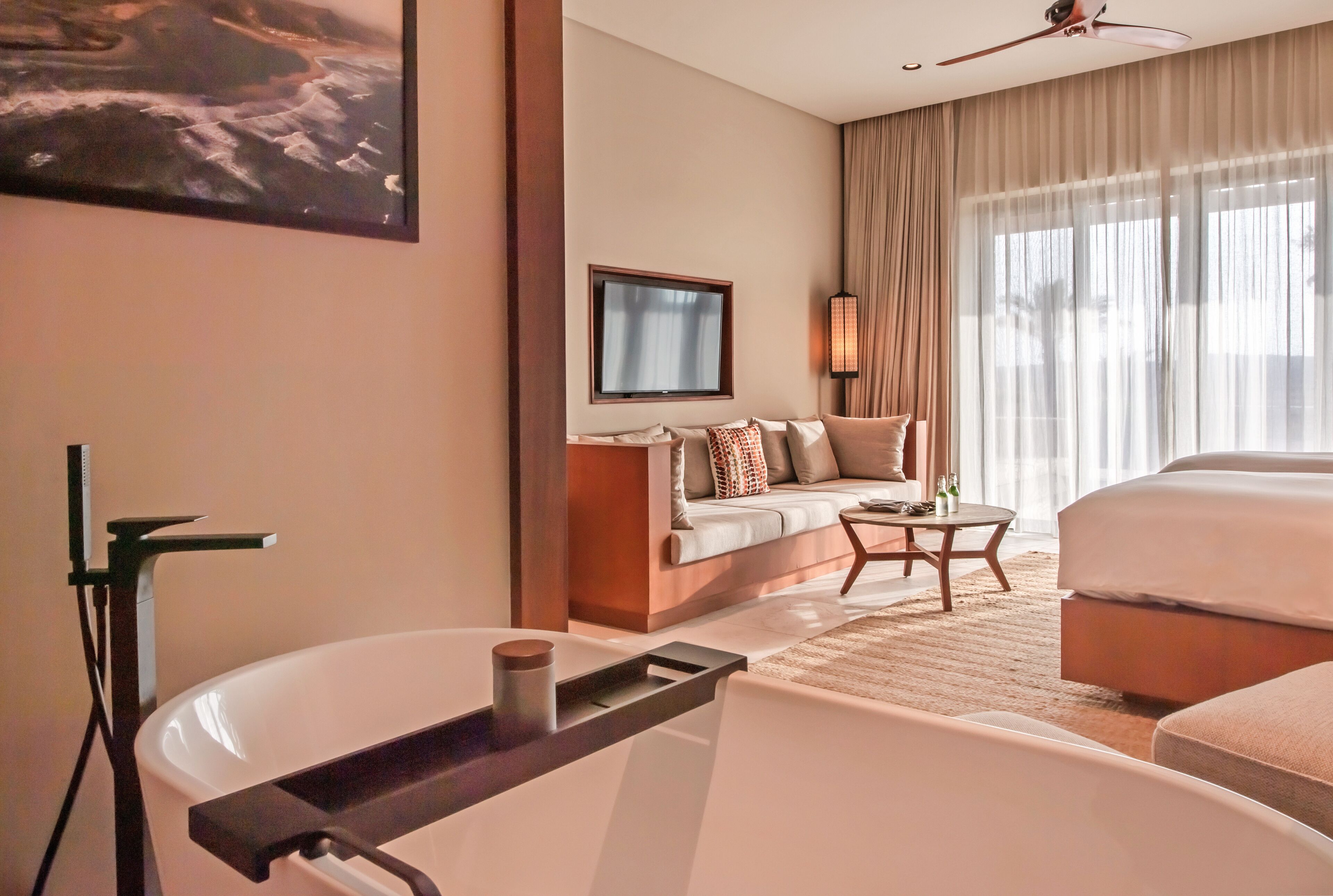 A modern hotel room with a large bathtub, a sofa, a television, and a large window with curtains.
