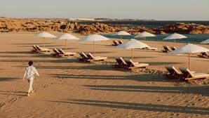A beach with rows of sun loungers and umbrellas, a person walking, and a rocky coastline in the background. - Alila Hinu Bay (Mirbat)