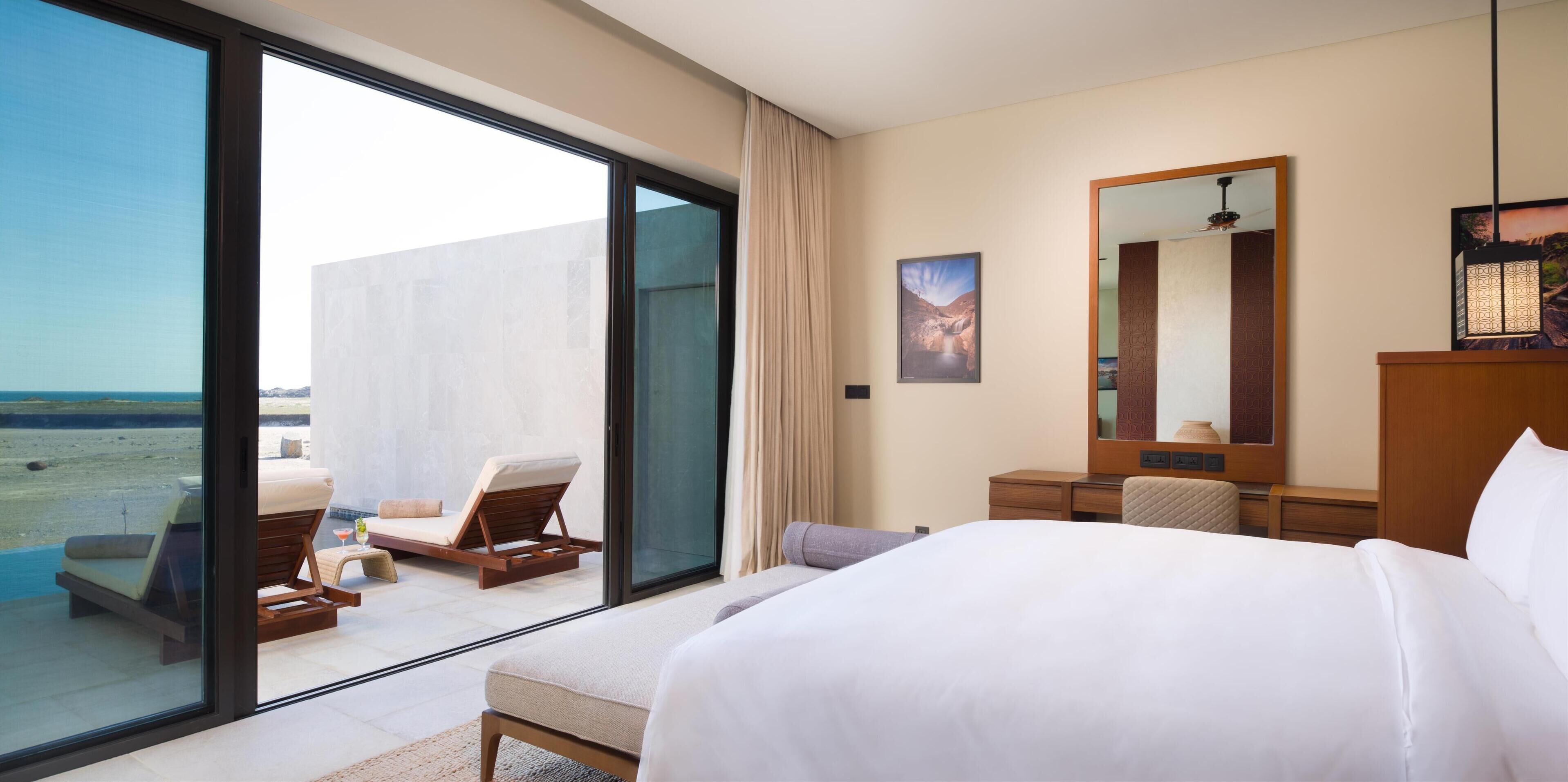 A hotel room with a large bed, a desk, a chair, a small table with a drink, and a large window offering a view of the beach.