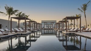 A pool area with lounge chairs and umbrellas, surrounded by palm trees and modern buildings. - Alila Hinu Bay (Mirbat)
