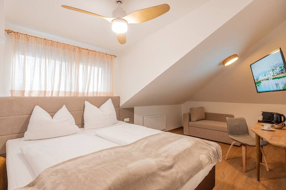 A bedroom with a bed, a ceiling fan, a window with curtains, a radiator, a sofa, a chair, a table with a coffee maker, and a TV mounted on the wall.