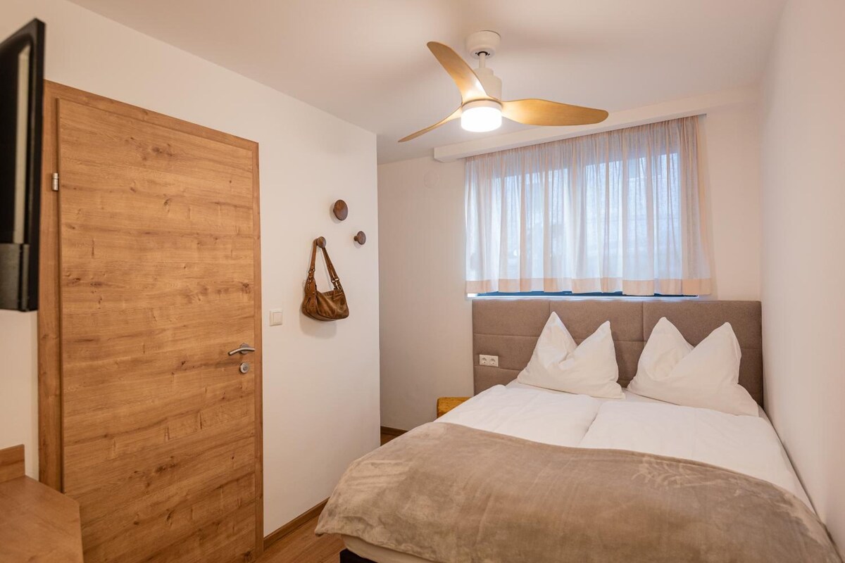 A bedroom with a wooden door, a ceiling fan, a window with sheer curtains, and a bed with multiple pillows.