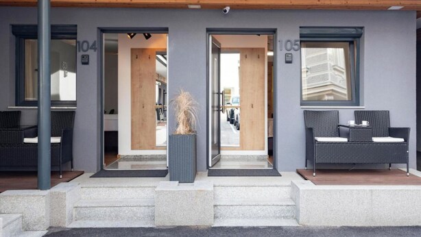 Entrada de hotel con dos puertas identificadas con los números 104 y 105, un pequeño espacio exterior con mobiliario de mimbre y una planta en maceta en el centro.