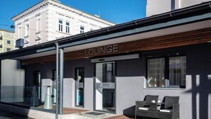 Un edificio moderno con un letrero que dice 'LOUNGE', que cuenta con una entrada de puerta de cristal, un pequeño espacio de estar al aire libre con sillas de mimbre y una terraza de madera.