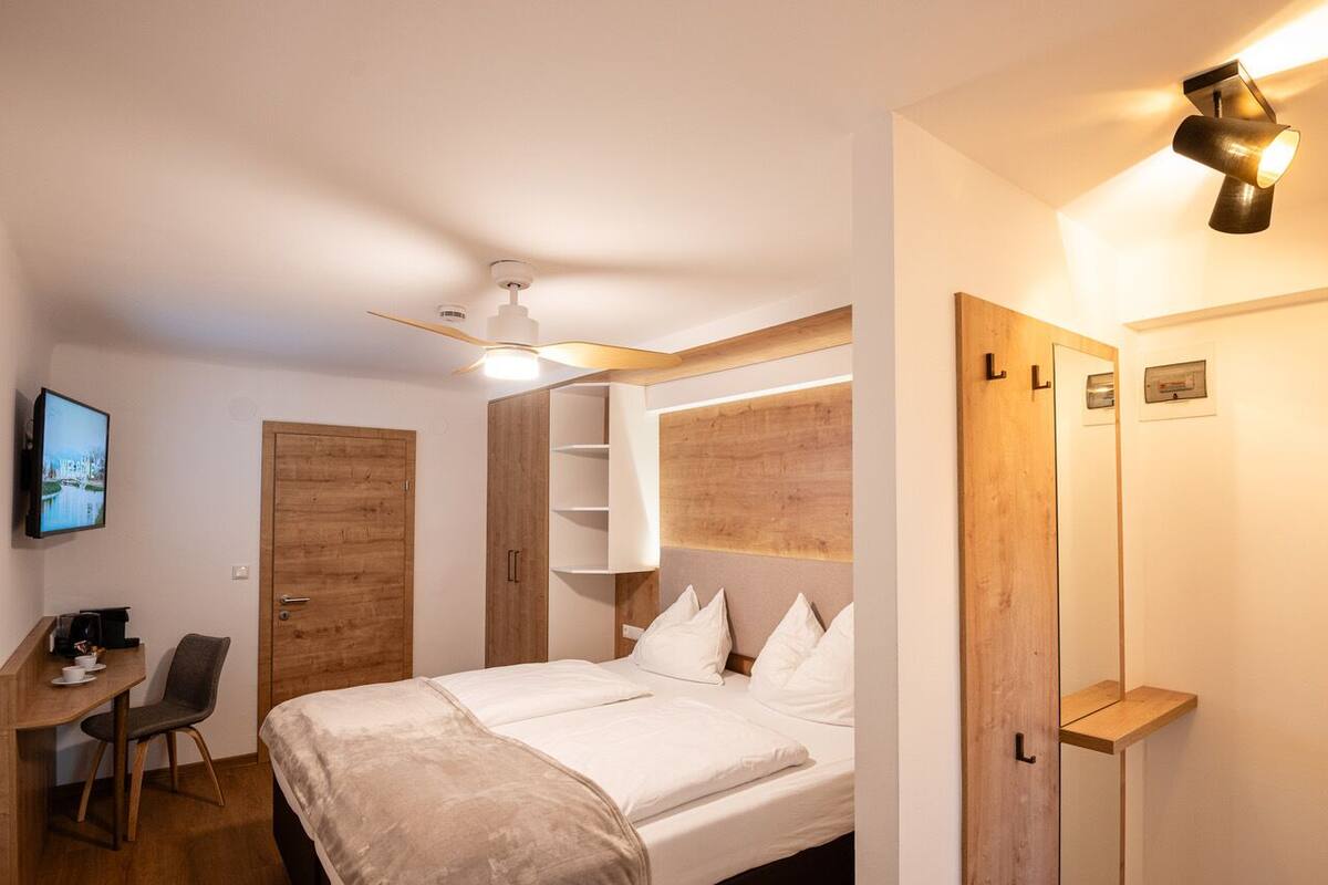 A modern hotel room with a bed, a desk with a chair, a TV, and a ceiling fan.