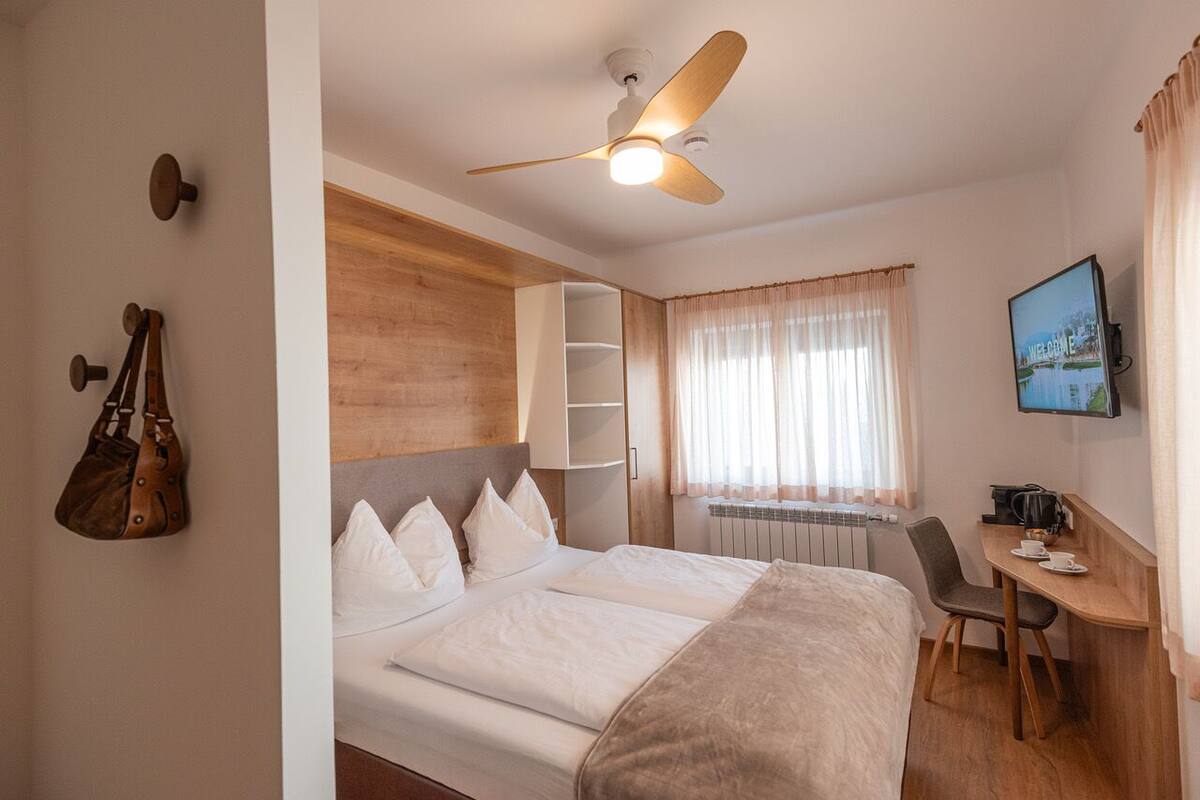 A hotel room with a bed, a desk, a chair, a ceiling fan, a wall-mounted TV, and a window with curtains.