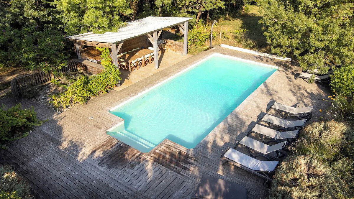 Une piscine rectangulaire entourée d’une terrasse en bois, avec un espace salon couvert et des chaises longues.
