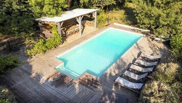 Une piscine rectangulaire entourée d’une terrasse en bois, avec un espace salon couvert et des chaises longues.