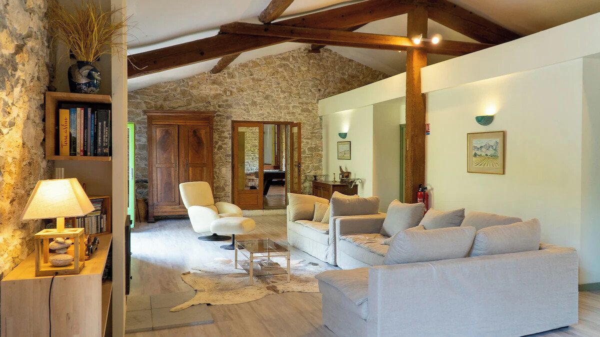 Un intérieur rustique avec un mur en pierre, des poutres en bois, un canapé blanc, un fauteuil et une petite table avec une lampe.