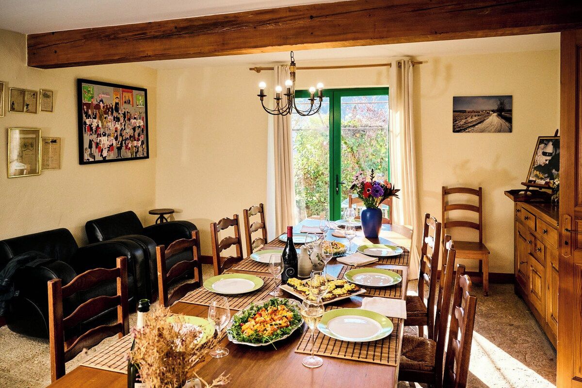 A dining area with a wooden table set for a meal, surrounded by chairs, with a view of greenery through a green-framed window.