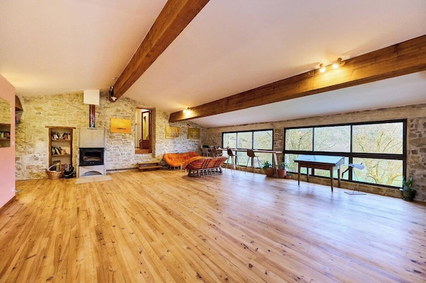A spacious room with a stone wall, wooden beams, a fireplace, and large windows.