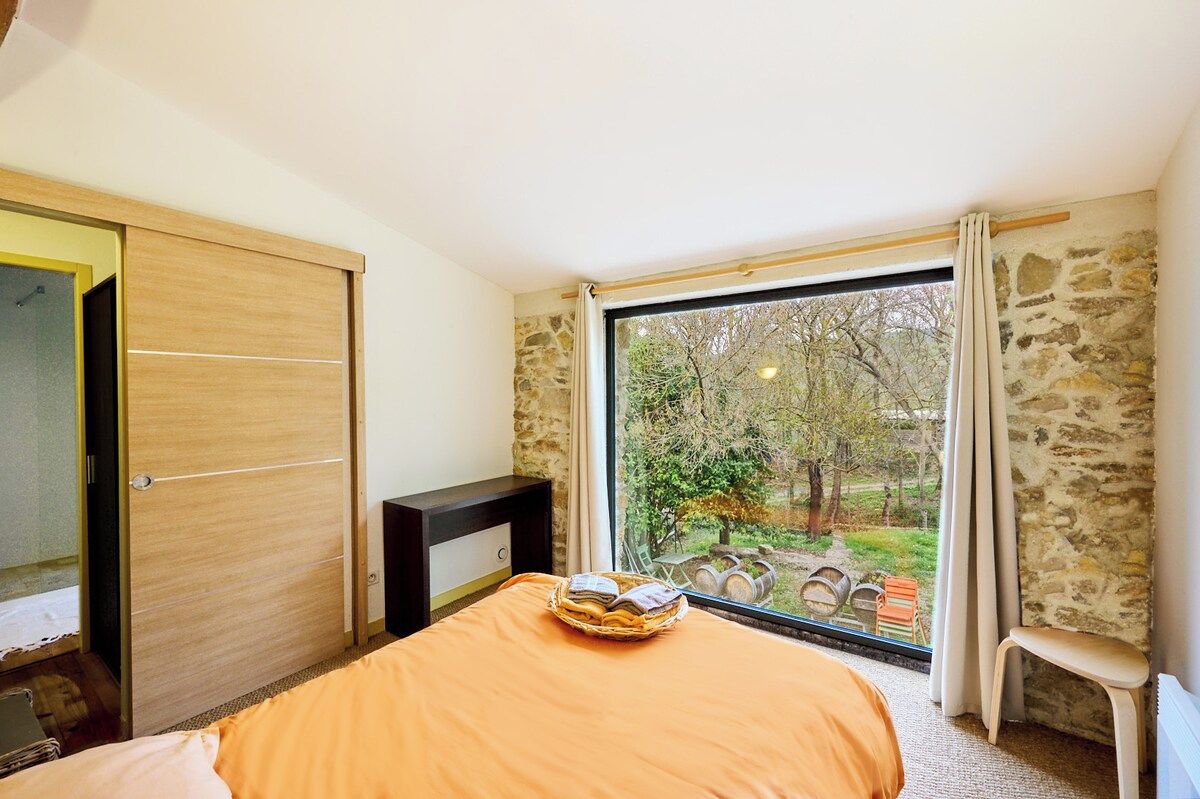 Een slaapkamer met een bed, een houten garderobe, een nachtkastje en een raam met uitzicht op bomen en vaten buiten.