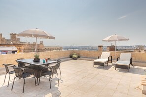 Una terraza en la azotea con una mesa, sillas, dos sombrillas y plantas en macetas.