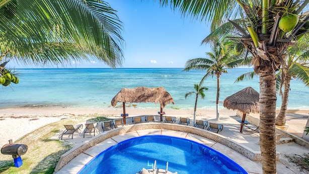 Ein Swimmingpool mit Steinkante, umgeben von Liegestühlen und einem Strohdachschirm, mit Blick auf einen Strand mit Palmen und dem Meer.