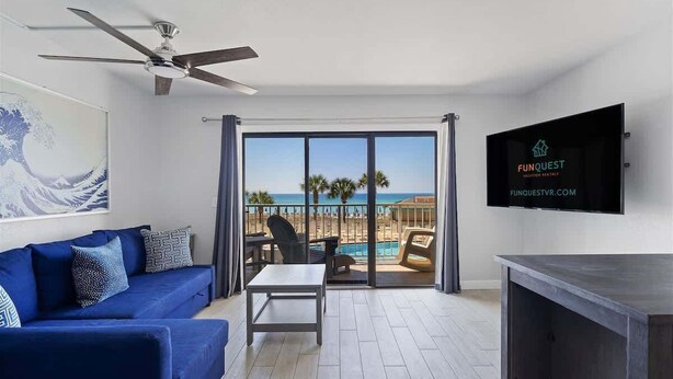 En moderne stue med en blå sofa, et lille bord og en ventilator i loftet. Et fjernsyn er monteret på væggen, og et stort vindue giver udsigt over stranden og havet.