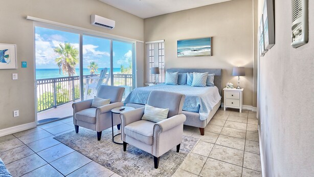 A bedroom with a large bed, a ceiling fan, a nightstand, and a view of the ocean through a sliding door.