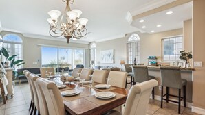 A spacious dining area with a wooden table set for six, surrounded by beige upholstered chairs. A chandelier hangs from the ceiling, and the room features large windows with a view of the outdoors.