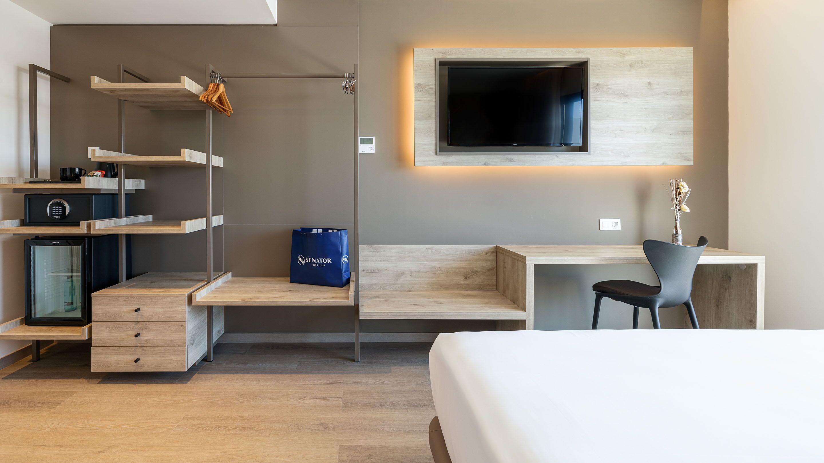 Una moderna camera d'albergo con una scrivania in legno, una sedia, una TV a parete, un minibar e un frigorifero per il minibar.