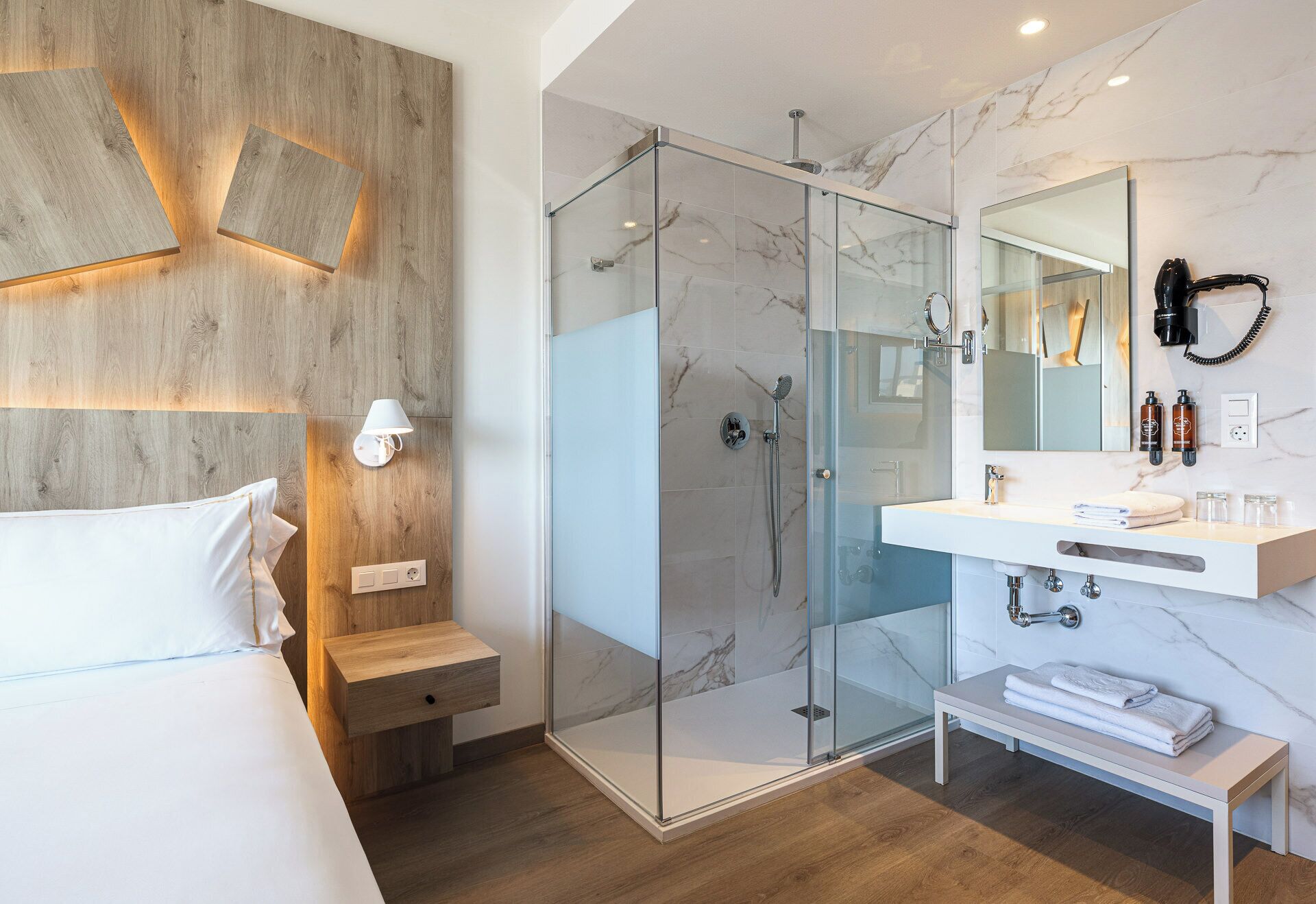 Une chambre d’hôtel moderne dotée d’une douche en verre, d’un mur d’accent en bois, d’une table de chevet avec une lampe et d’une salle de bain avec un comptoir en marbre et un grand miroir.