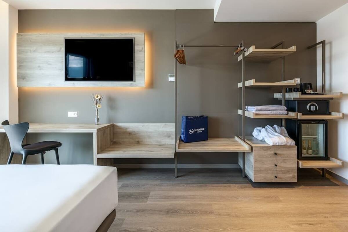 Une chambre d’hôtel moderne avec un bureau en bois, un lit, un téléviseur fixé au mur et des rangements intégrés.