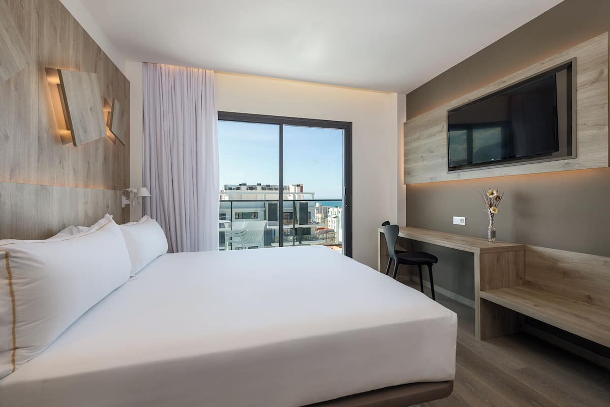 Une chambre d’hôtel moderne dotée d’un grand lit, d’un bureau avec une chaise, d’un téléviseur fixé au mur et offrant une vue sur la ville grâce à une grande fenêtre.
