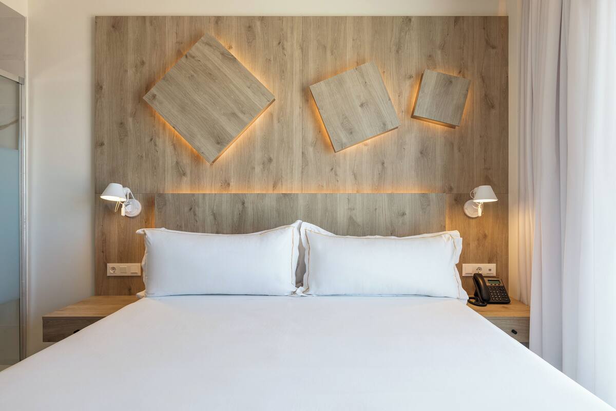 Une chambre d’hôtel moderne avec une tête de lit en bois, deux lampes de chevet, un téléphone et un lit parfaitement fait.