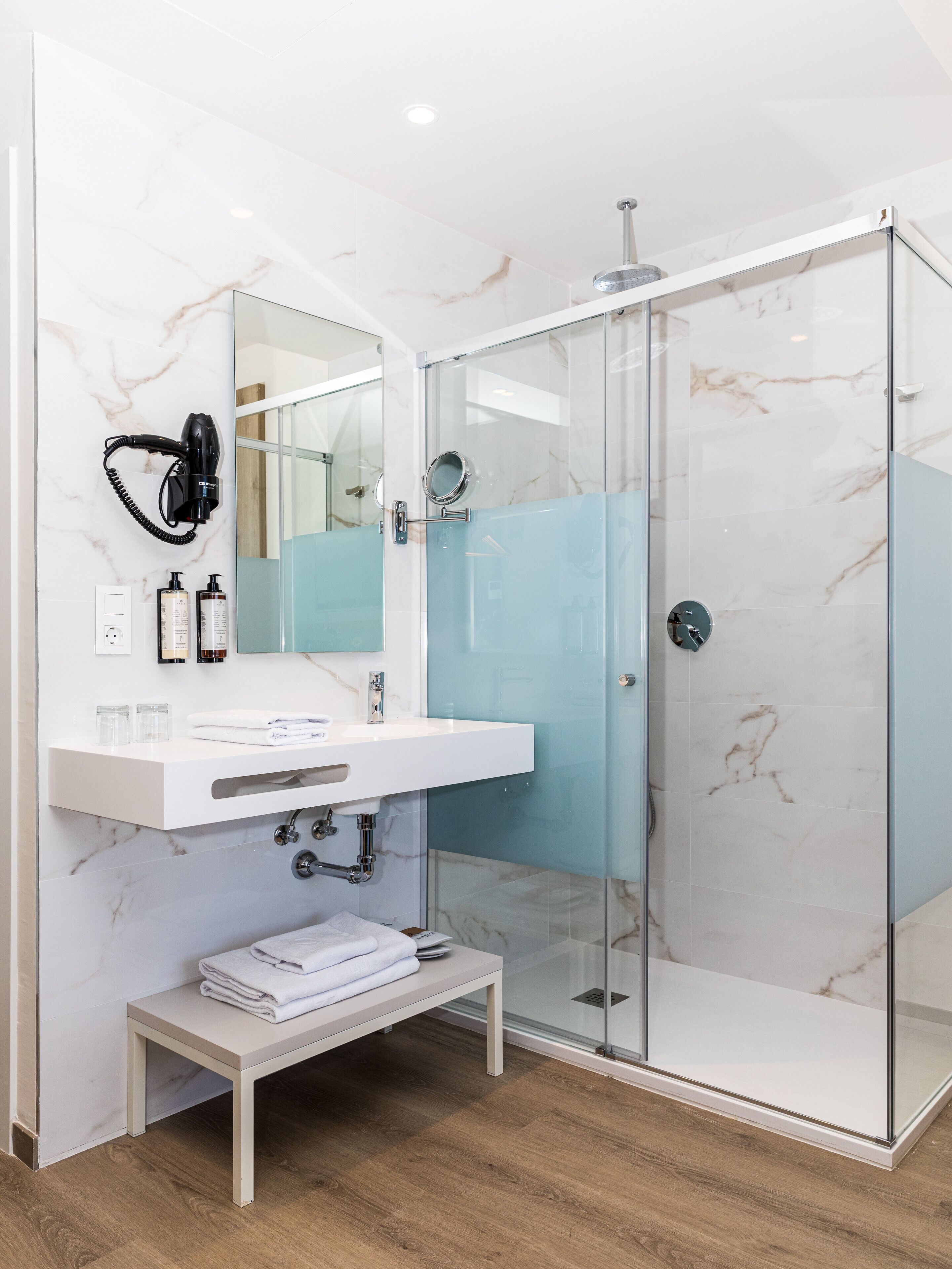 A modern bathroom with a glass shower enclosure, a white marble sink, and a hairdryer mounted on the wall.